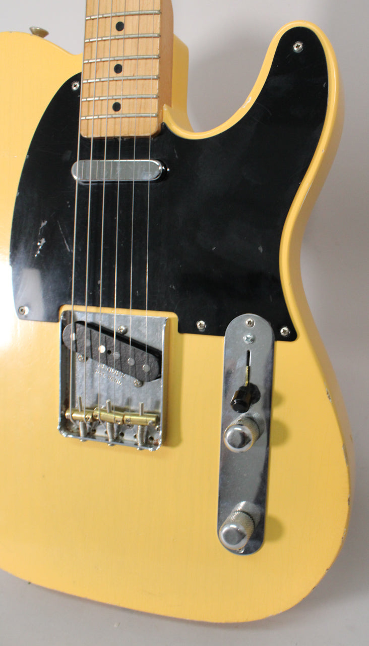 Close-up of a blonde Telecaster body with a black pickguard, showing a single-coil bridge pickup, three brass barrel saddles, and two chrome knobs.