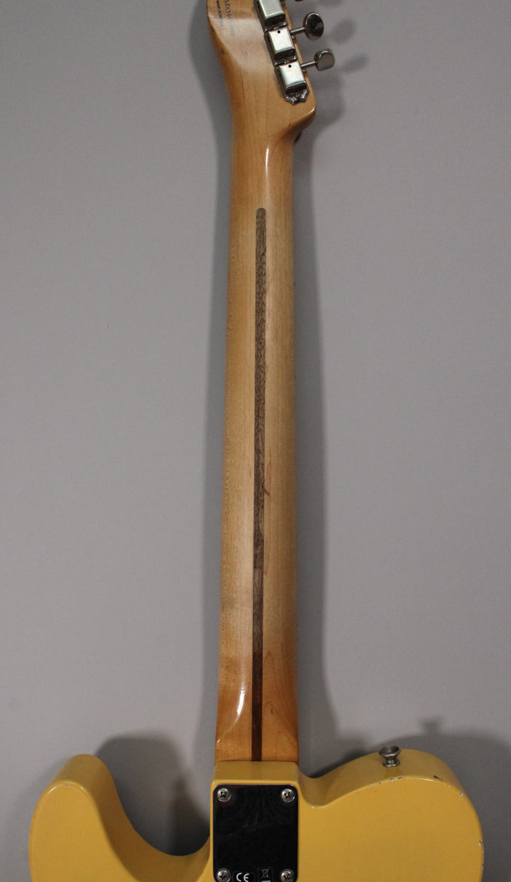 The guitar's blonde body and neck are viewed from the back, showing the light wood neck with a dark skunk stripe and four chrome tuners.