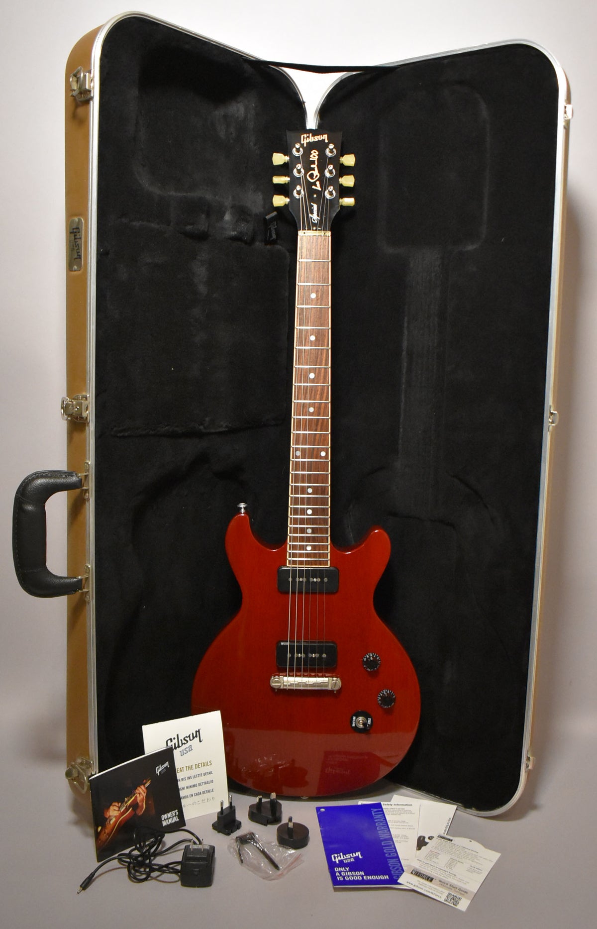 Full-length view of a cherry red double-cutaway electric guitar with two black P90 pickups, in a tan hard case with paperwork.
