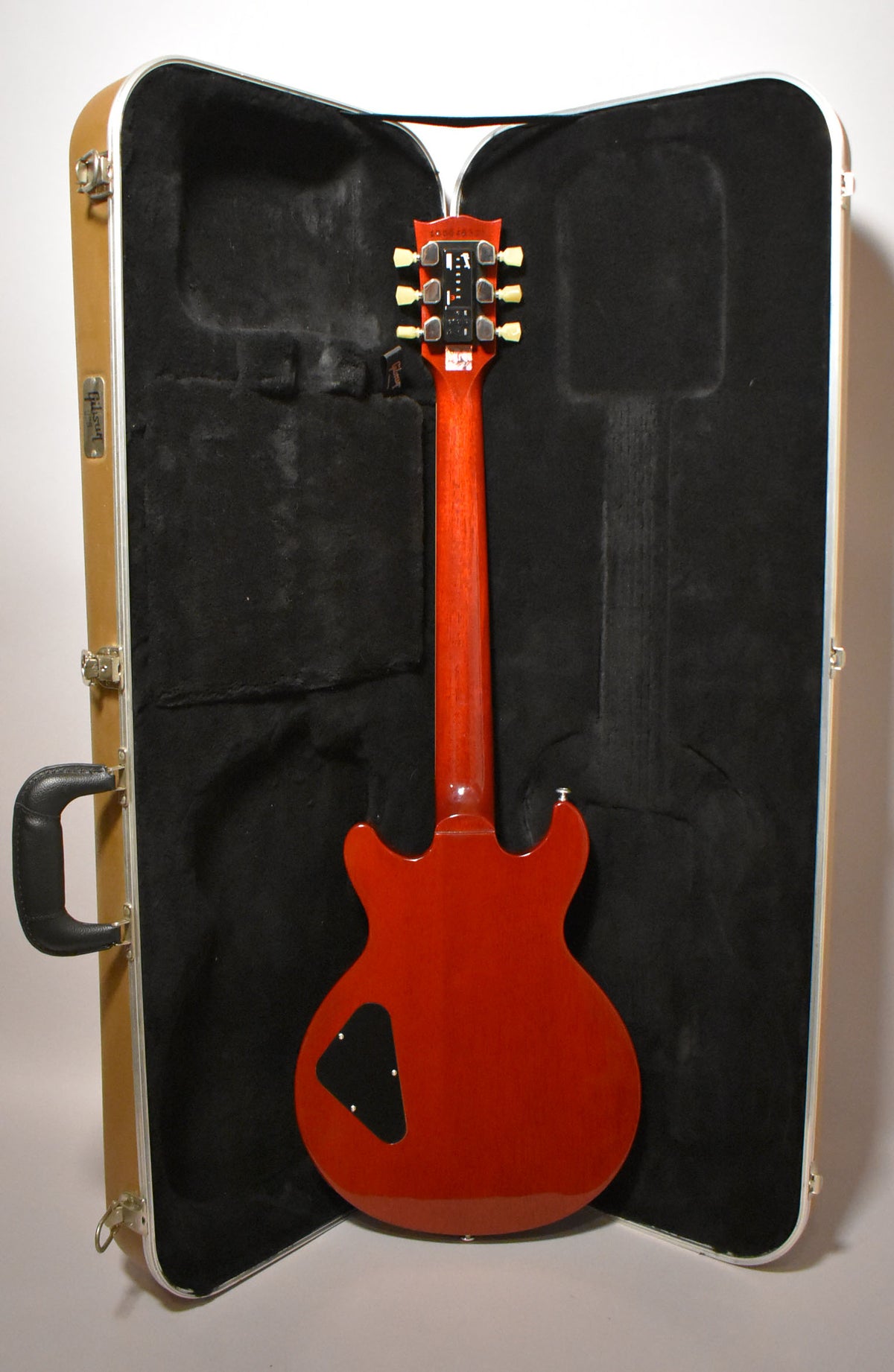 The cherry red guitar is shown from the back, resting in its open, tan hard case with a black plush interior.