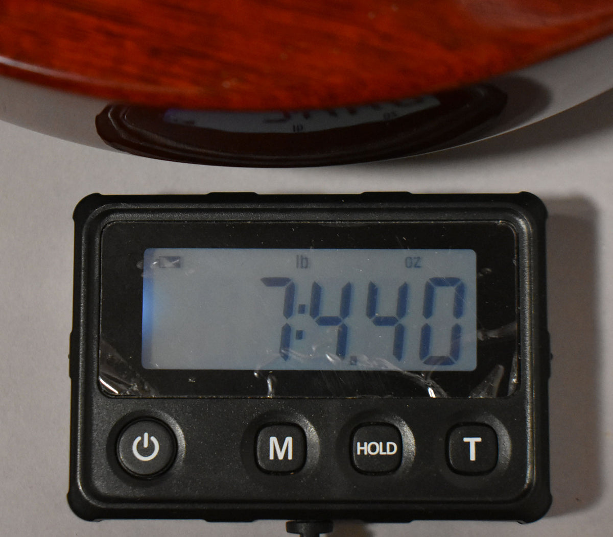 Close-up of a digital scale displaying "7:44.0 lb oz", with a cherry red guitar body resting on the scale's platform.