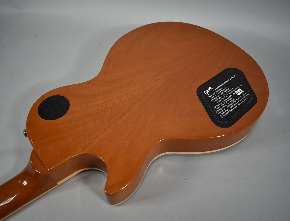 The guitar's glossy brown back is angled, revealing a black circular control plate and a rectangular sticker with white text.