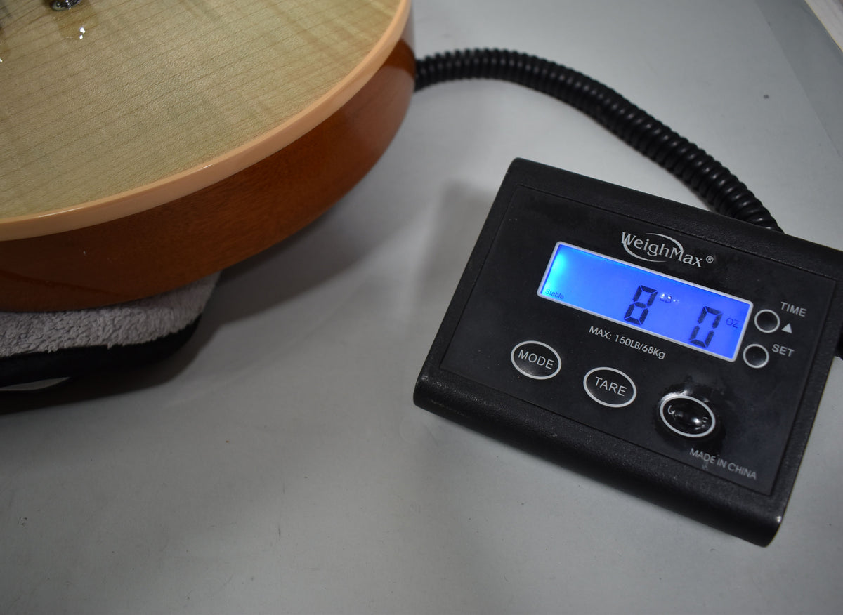 The guitar's light-colored flamed maple top and brown back are visible, resting on a scale displaying "8 lb 0 oz".