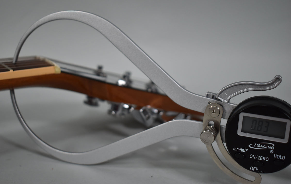 A digital caliper measures the guitar's neck thickness at the nut, displaying 0.83 inches, with tuning pegs visible behind.