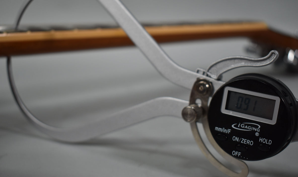 A digital caliper measures the neck thickness at 0.91 inches, with the guitar's light brown neck out of focus behind it.