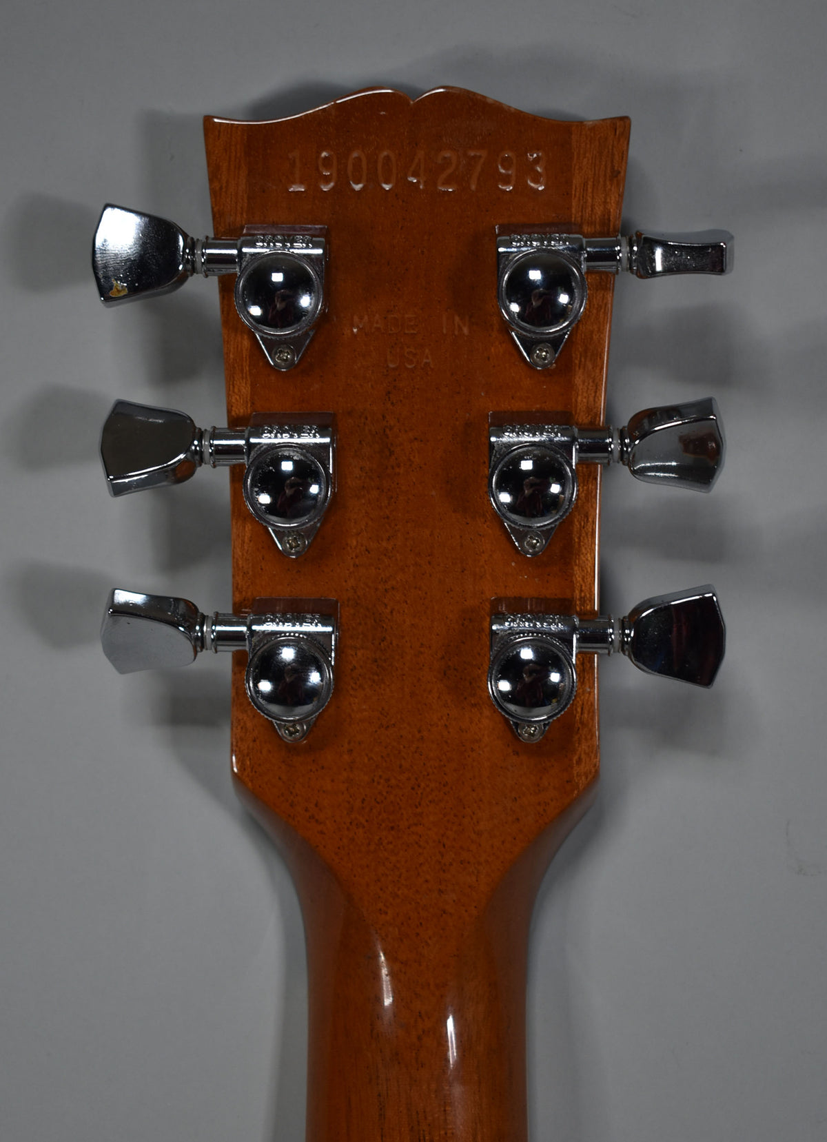 Rear view of the guitar's headstock and neck, showing a glossy brown finish, six chrome tuners, and a serial number.