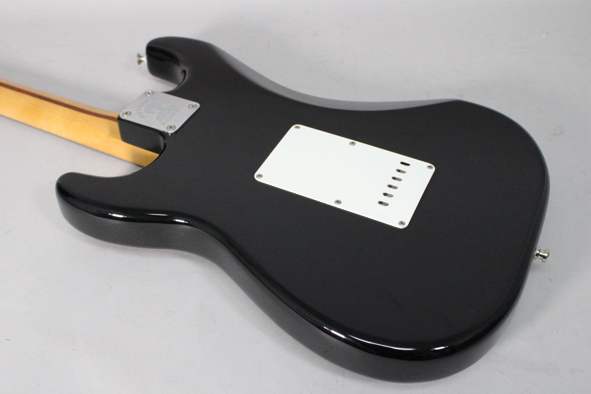The back of a glossy black Stratocaster body is angled, showing a white tremolo cover, chrome neck plate, and light wood neck.