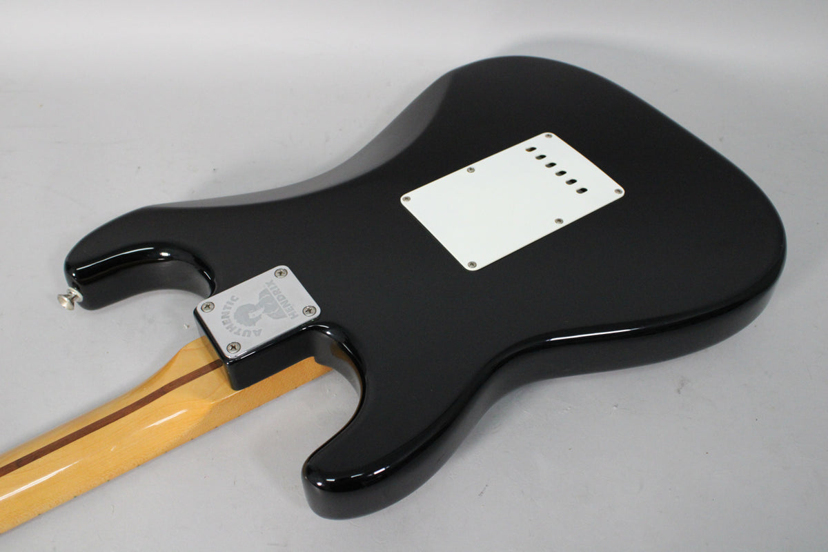 The guitar's glossy black back is angled, showing a white tremolo cover and a chrome neck plate with a circular design.
