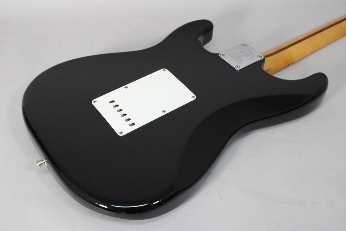 The black Stratocaster's glossy back shows a white tremolo cover, chrome neck plate, and a light maple neck with a dark stripe.