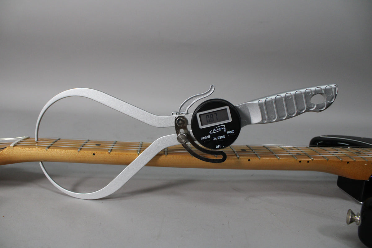 A digital caliper measures the neck thickness of the black Stratocaster, displaying "0.87" on its screen.