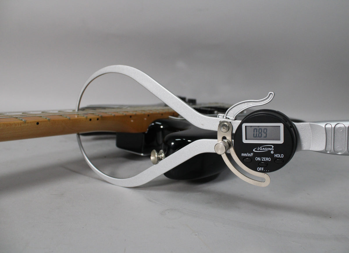 A digital caliper measures the neck relief of the black Stratocaster, showing 0.89mm, with the maple fretboard visible.