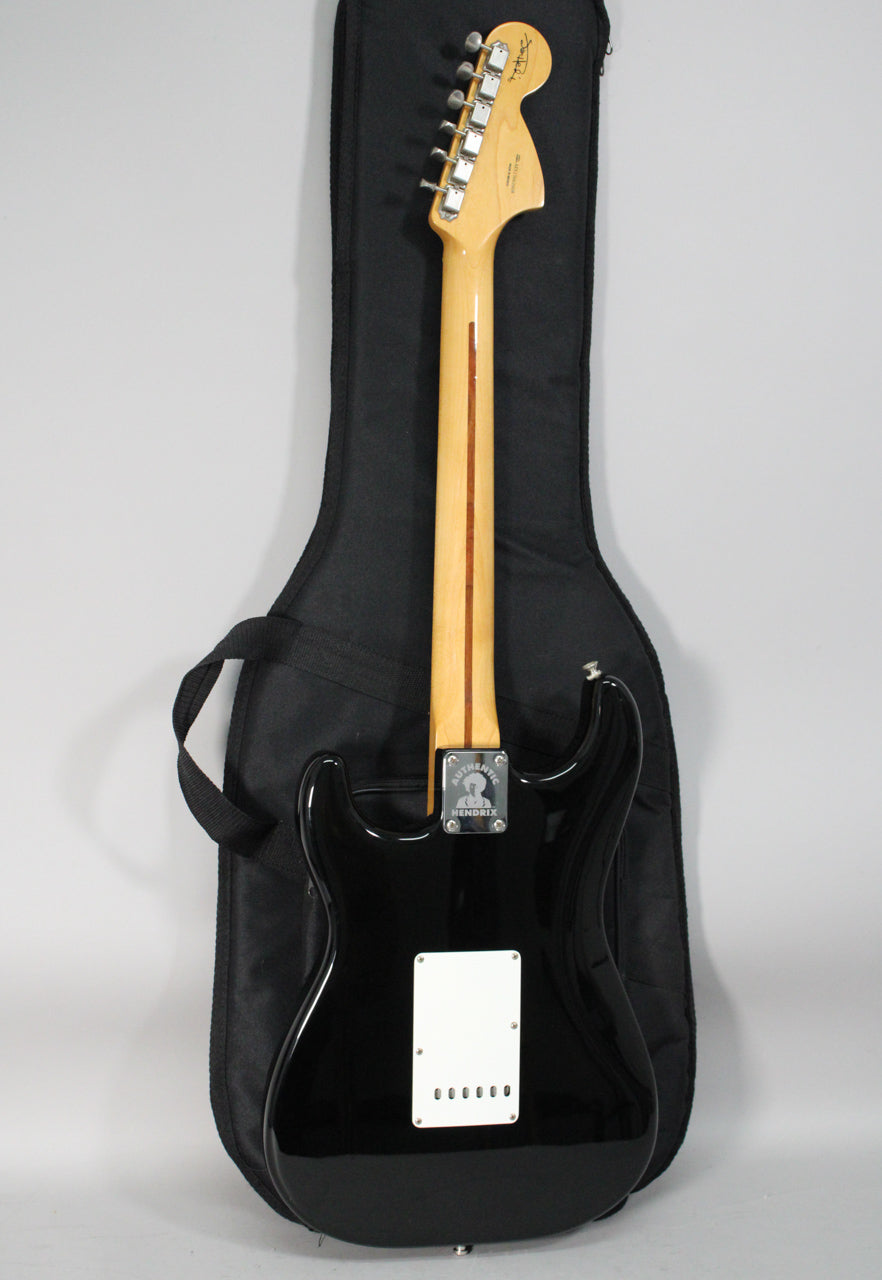 The back of a glossy black Stratocaster with a light maple neck, chrome tuners, and a white tremolo cover, resting in a black gig bag.