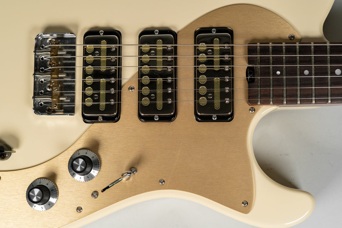 Close-up of an antique white guitar body with a brushed gold pickguard, three gold foil pickups, a chrome bridge, and two chrome knobs.