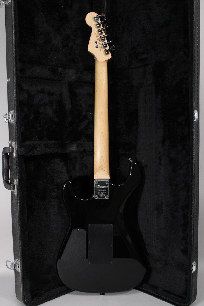 The guitar's black body and light maple neck are seen from the back, resting in a black plush-lined hard case.