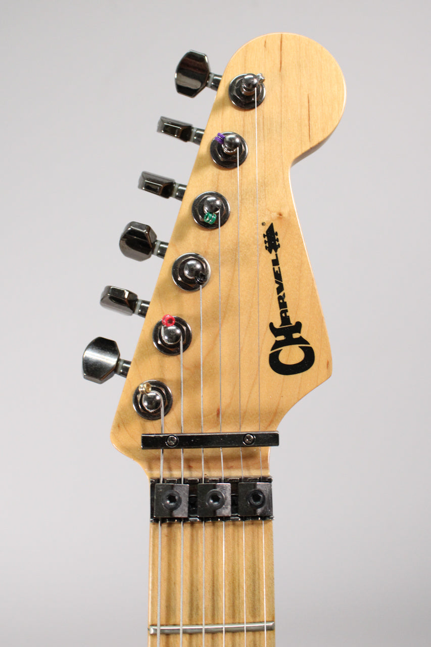 Close-up of a guitar headstock with a natural wood finish, black Charvel logo, six chrome tuners, and a black locking nut.