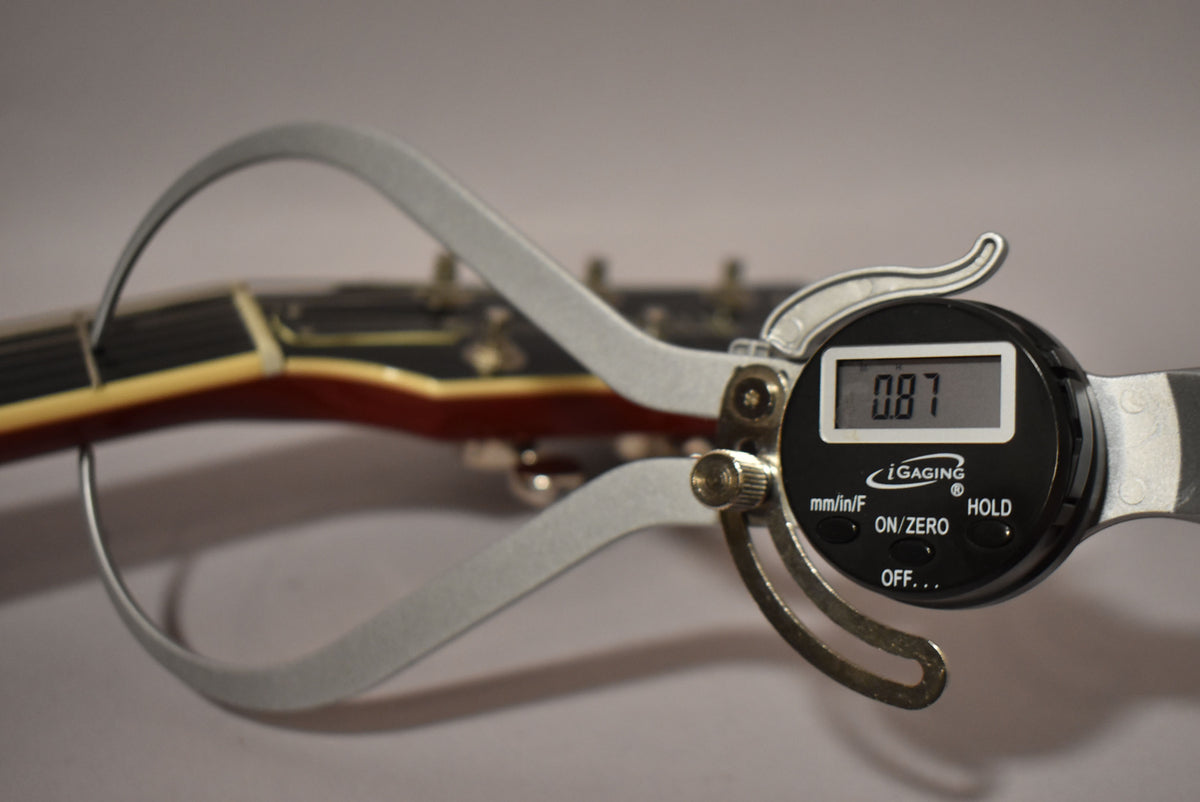 A close-up of the guitar's headstock and fretboard, with a digital caliper measuring the fret height at 0.87.