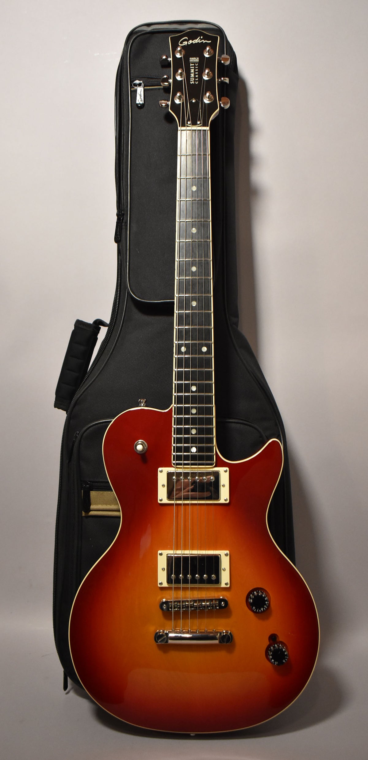 Full-length view of a cherry burst electric guitar with a glossy finish, cream binding, two humbuckers, and a black gig bag behind it.