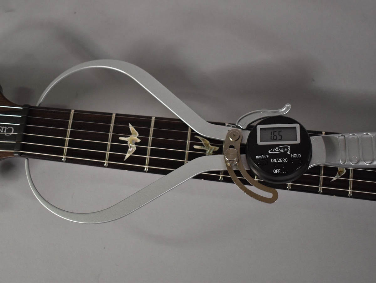 Close-up of the dark fretboard with bird inlays, showing a digital caliper measuring 1.65 inches across the nut.