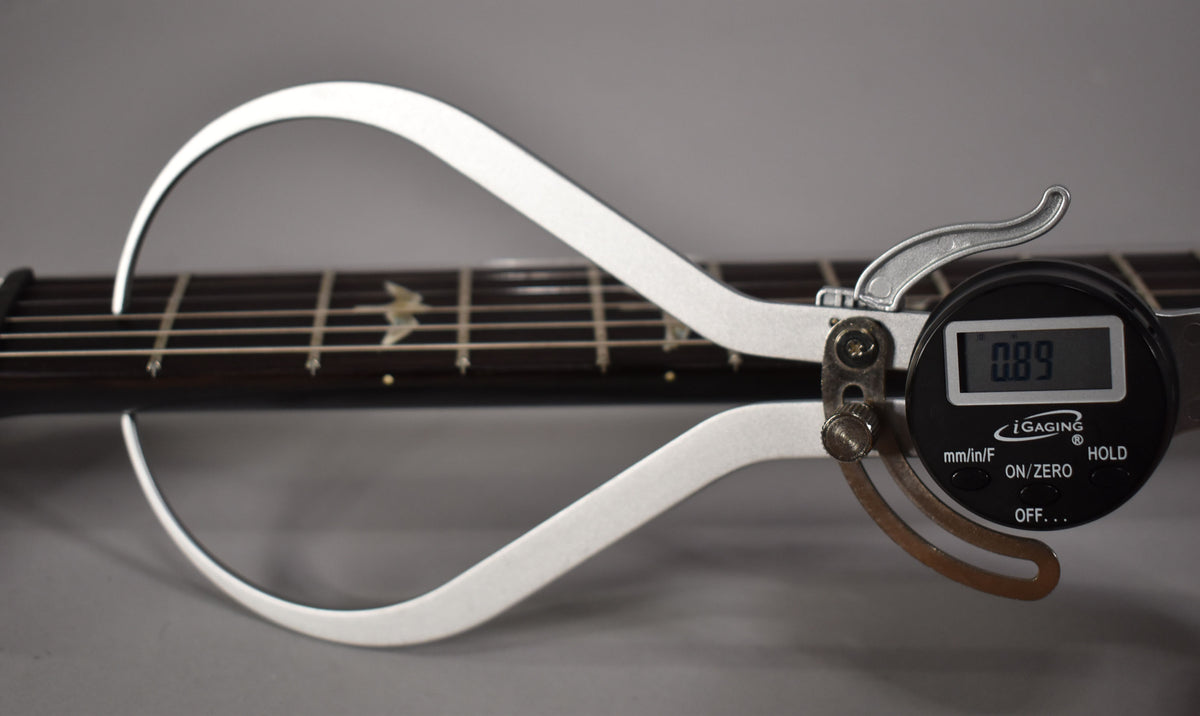 A digital fret height gauge measures the guitar's dark fretboard with bird inlays and light strings.