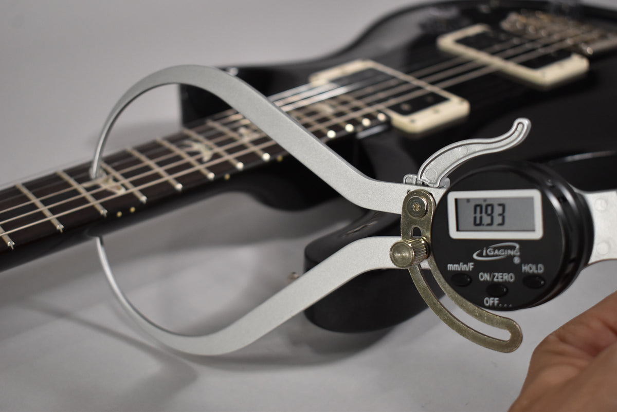 A digital caliper measures the fret height at 0.93mm on the black guitar's fretboard, with cream humbuckers visible in the background.