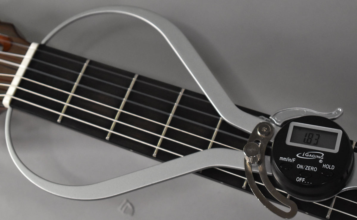 A digital fret height gauge, displaying "1.83", measures the frets on the dark fretboard of the classical guitar.