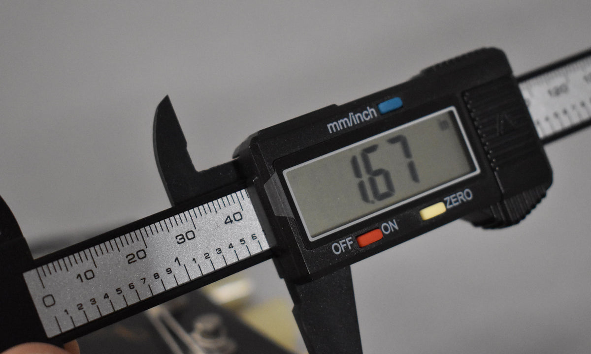 A digital caliper measures 1.67mm, clamped onto a guitar's nut, with strings visible behind the measuring jaws.