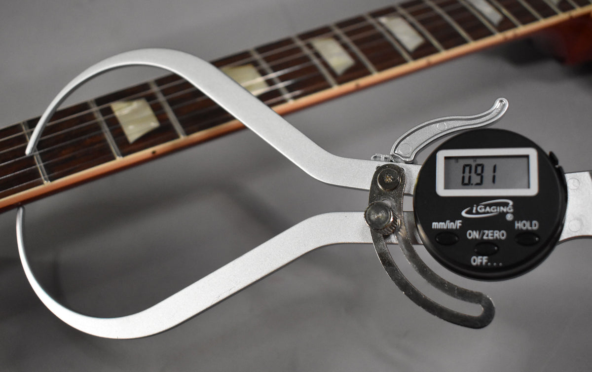 A digital caliper measures the guitar's neck thickness at 0.91 inches, with the dark fretboard and trapezoid inlays visible.