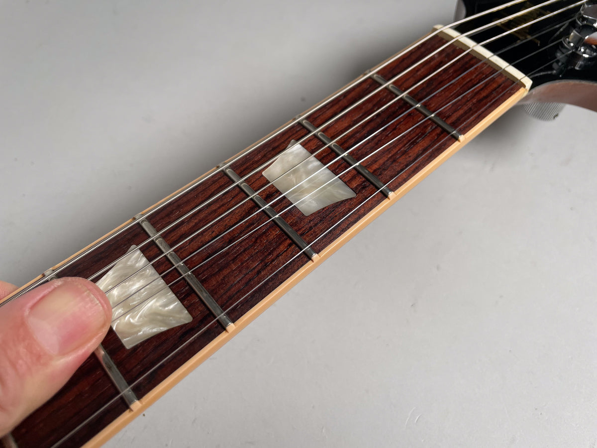 Close-up of the guitar's dark rosewood fretboard with trapezoid mother-of-pearl inlays, a thumb rests on the first inlay.