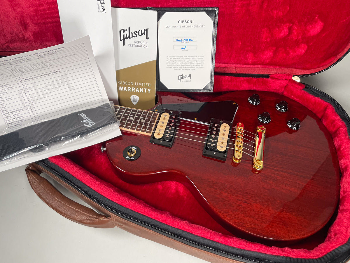 A cherry red electric guitar with gold hardware and black knobs, nestled in a red-lined case with paperwork and a strap.