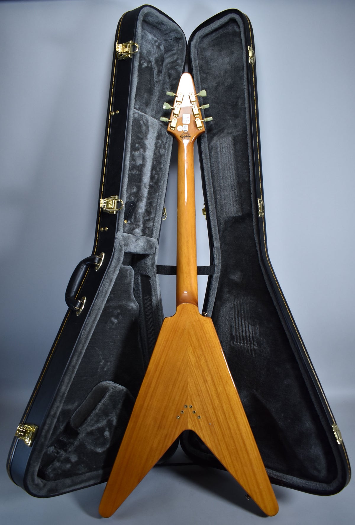 The natural finish Flying V guitar is seen from the back, standing in its open, black, plush-lined hard case.