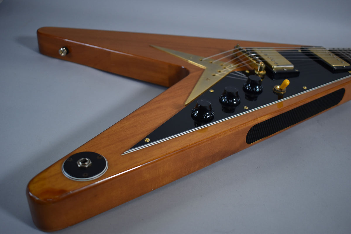 The lower half of a natural wood Flying V, showing the black pickguard, gold hardware, three black knobs, and yellow switch.