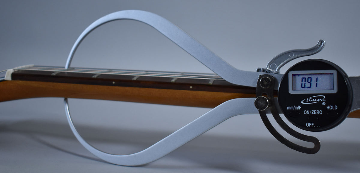 A digital caliper measures the guitar's natural finish neck and dark fretboard, displaying "0.91" on its screen.