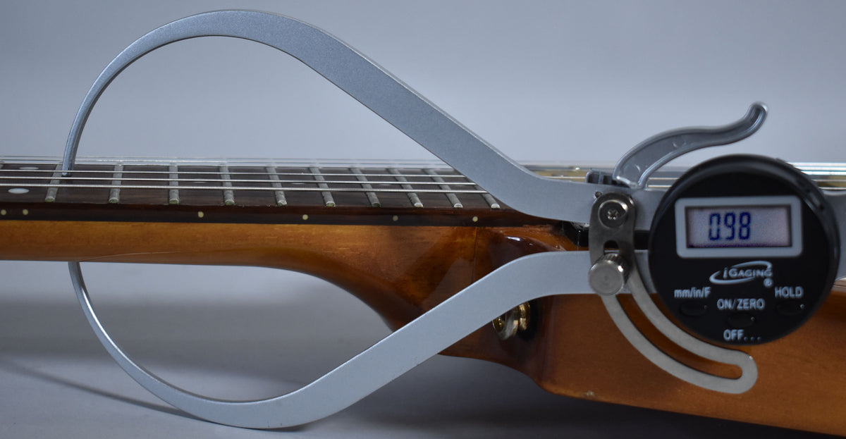 A digital caliper measures the neck relief of the natural finish guitar, showing a reading of 0.98.