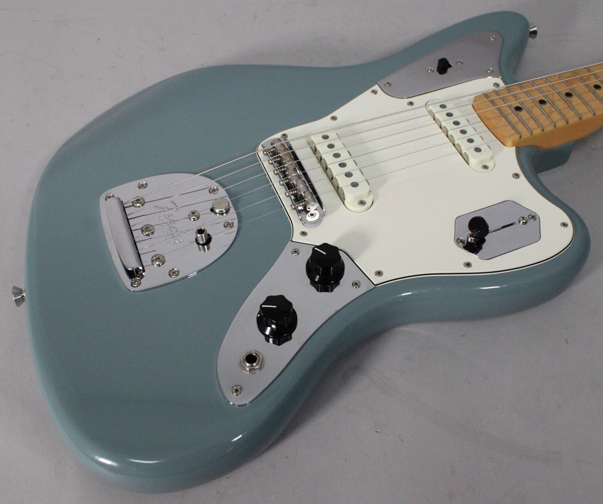 Light grey-blue Jaguar electric guitar, angled from above, showing the body, white pickguard, chrome hardware, and maple neck.