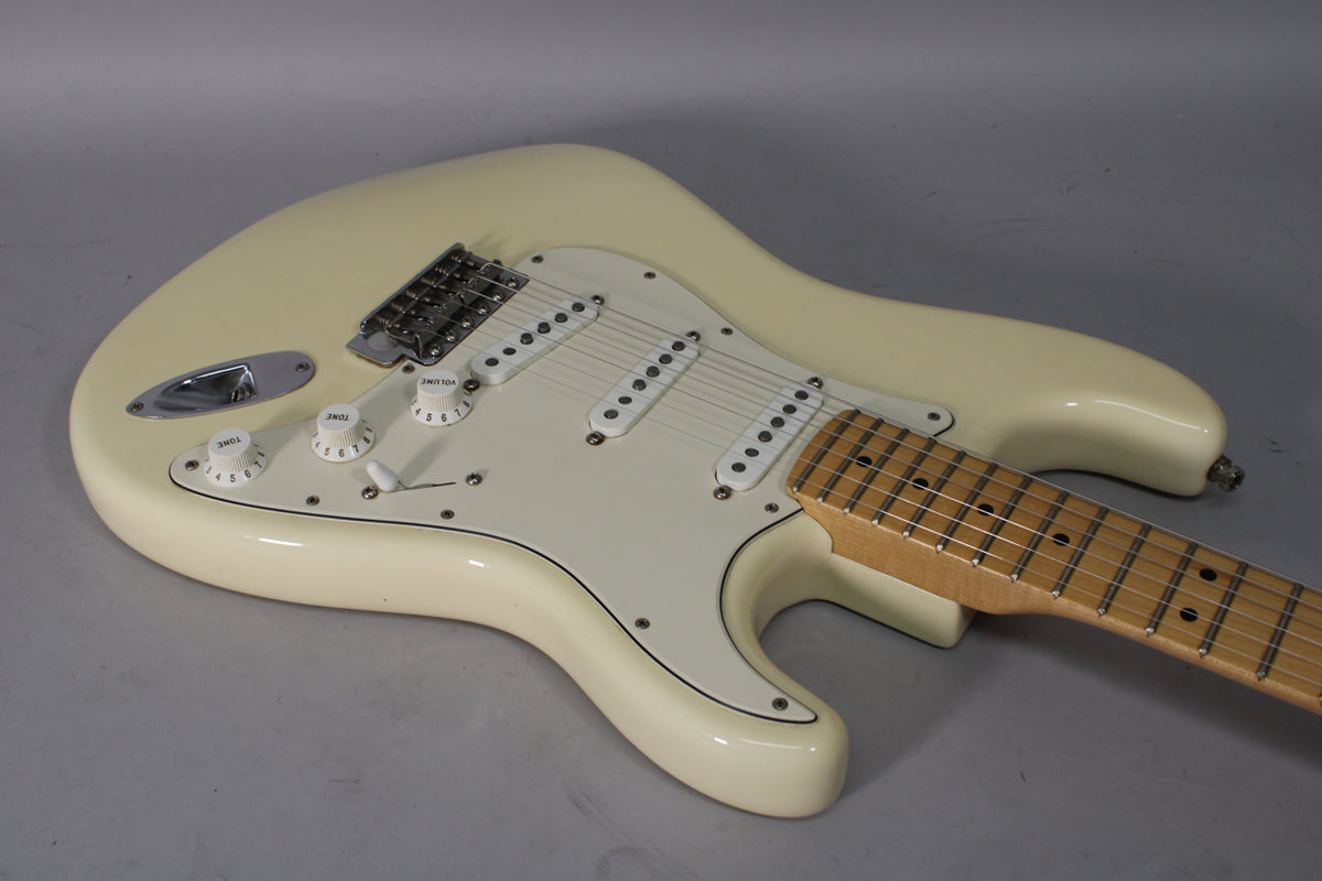 An Olympic White Stratocaster, angled from the lower left, showing the body, white pickguard, three single-coil pickups, and maple fretboard.