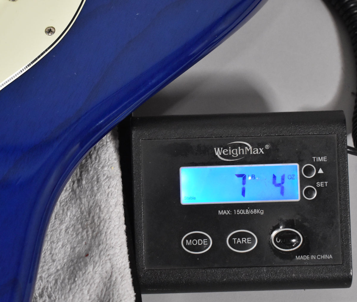The sapphire blue Stratocaster body rests on a white towel, with a digital scale displaying "7 LB. 4 OZ."