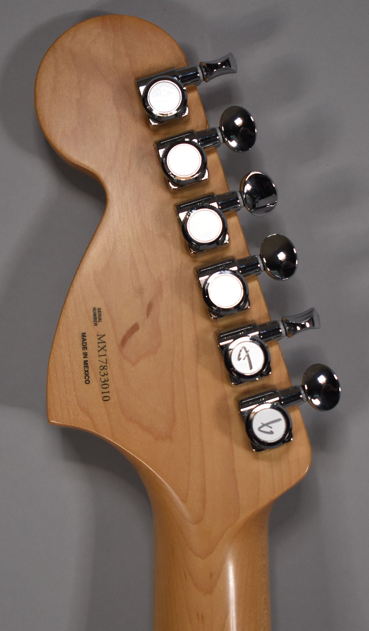 The back of a light maple headstock with six chrome tuning machines, showing a faint reddish stain and a serial number.