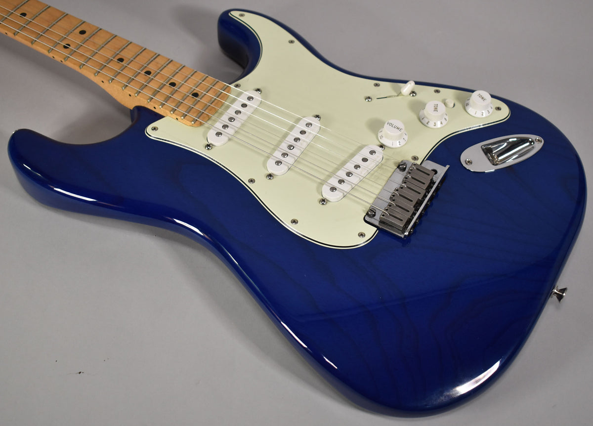 A sapphire blue electric guitar, angled from the upper left, shows its maple fretboard, cream pickguard, and three white single-coil pickups.