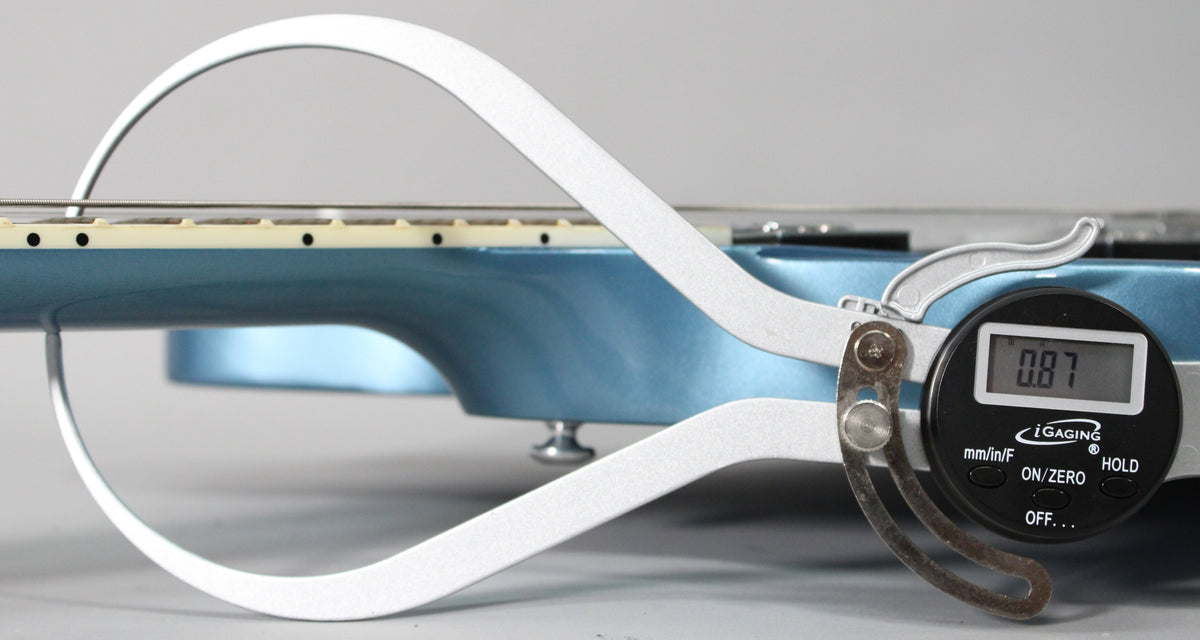 A digital string action gauge measures the Pelham Blue guitar's string height at 0.87mm, showing the fretboard and body.