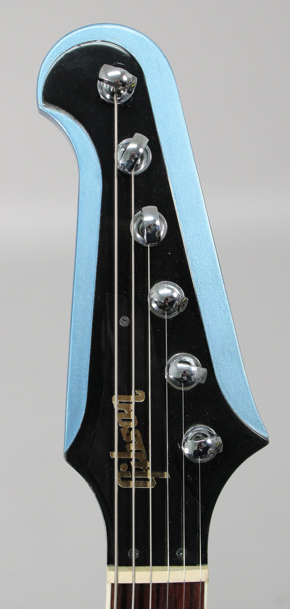 Close-up of the Pelham Blue Firebird's headstock, showing the black face with gold lettering, chrome tuners, and strings.