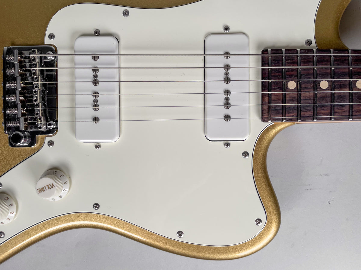 Close-up of a gold sparkle guitar body with a cream pickguard, two white S90 pickups, a chrome tremolo bridge, and two white control knobs.