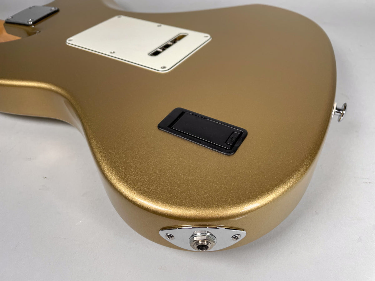 The back of a gold sparkle guitar body, showing the tremolo cavity cover, battery compartment, and input jack.