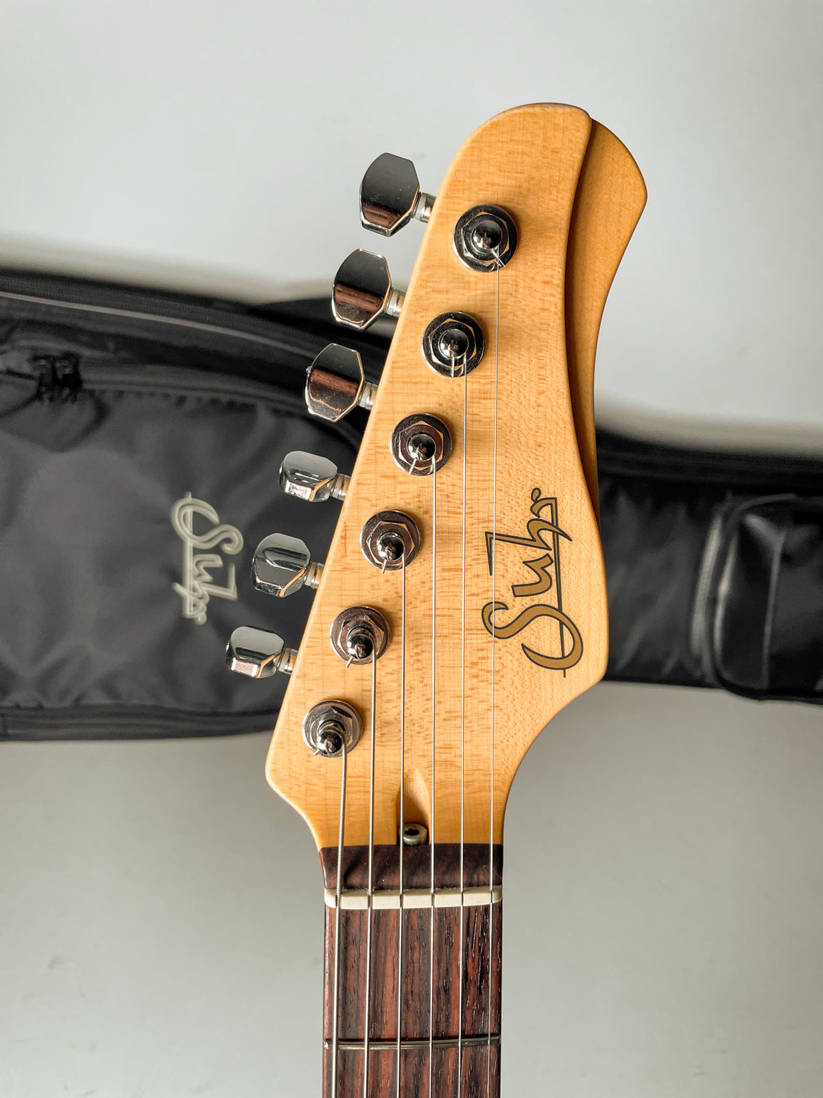 The guitar's light wood headstock with six chrome tuners and dark fretboard is angled slightly right, with a black gig bag behind.