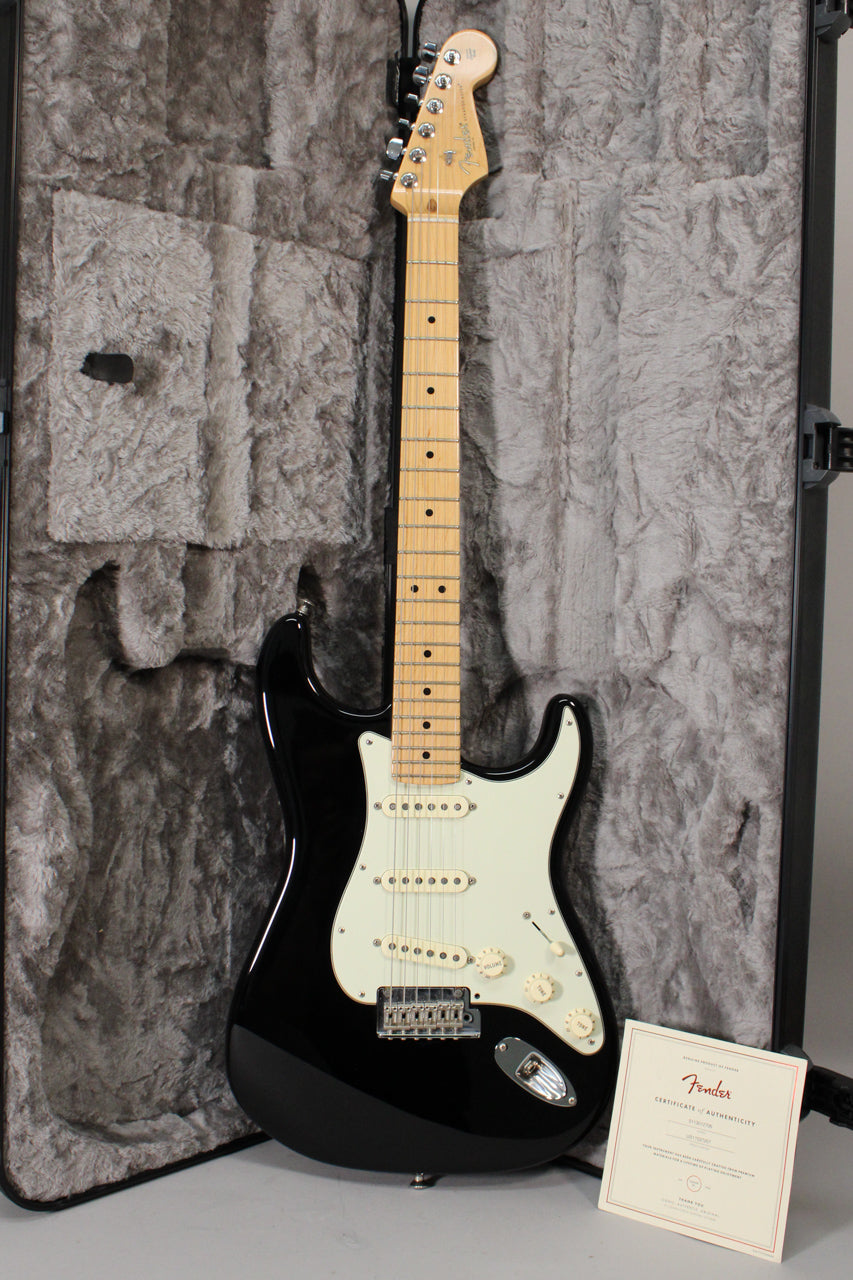 Full-length view of a glossy black Stratocaster with a maple neck, white pickguard, three single-coil pickups, and a tremolo bridge, standing in its open, grey-lined hard case with a certificate.