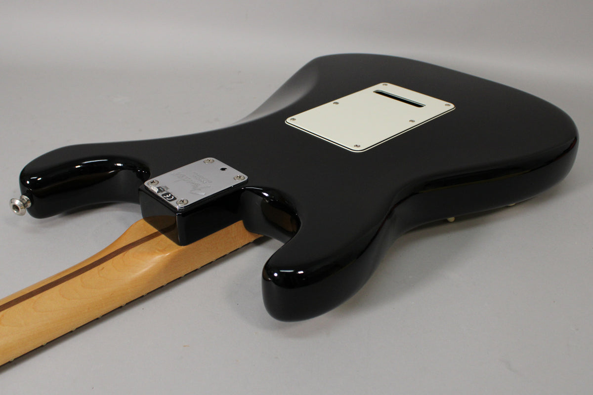 The glossy black guitar body is viewed from the back, showing the light maple neck, chrome neck plate, and white tremolo cover.