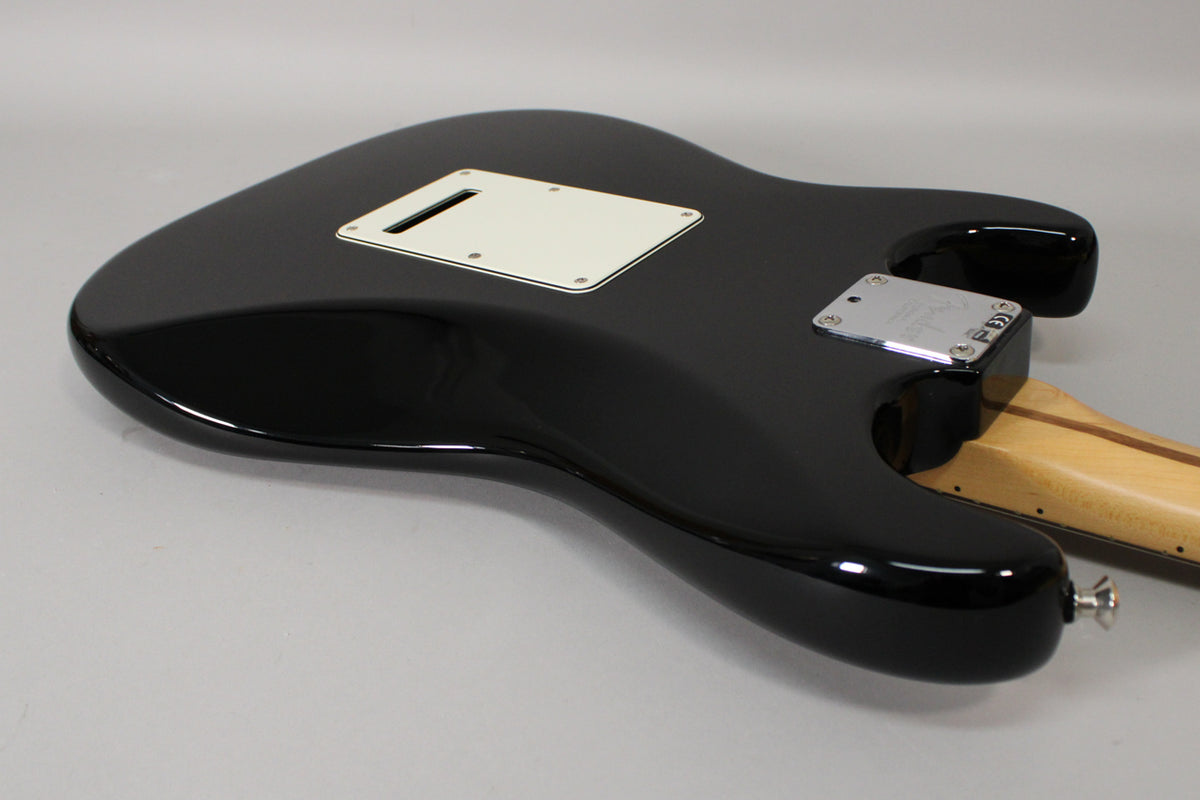 The black gloss Stratocaster body is viewed from the back, showing the white tremolo cover, chrome neck plate, and light maple neck.