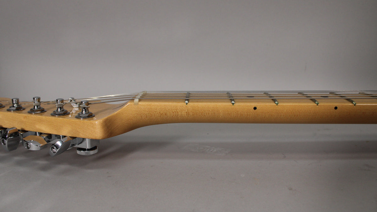 The guitar's light wood neck and headstock are viewed from the side, showing six chrome tuning machines and a few fret markers.