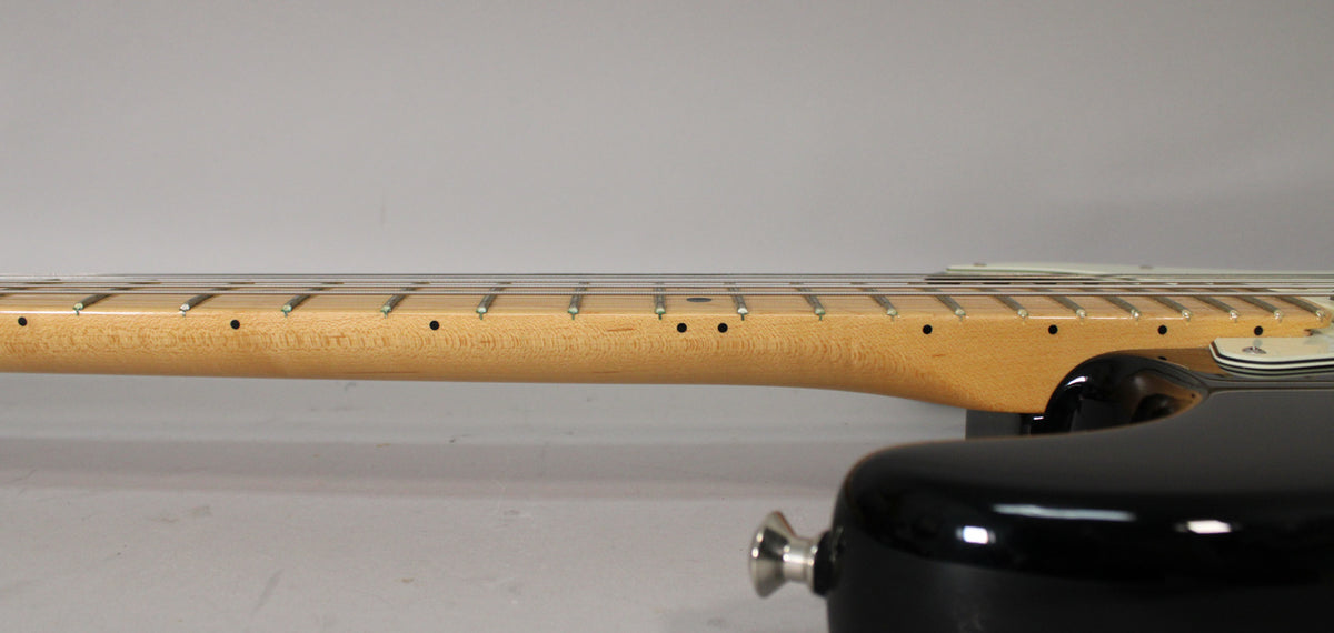 The black Stratocaster's maple neck and fretboard are shown from the side, with a chrome strap button visible on the body.