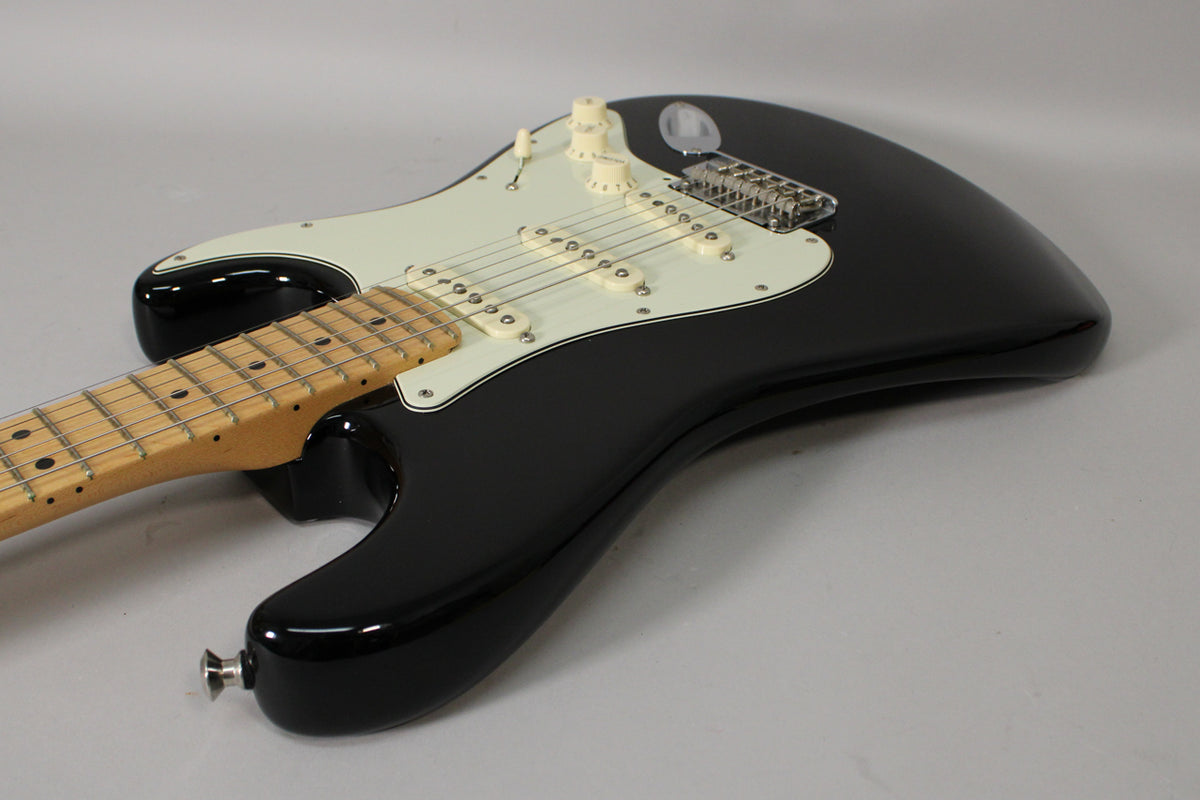 Black Stratocaster, angled from the lower bout, showing the glossy body, maple neck, white pickguard, and three single-coil pickups.