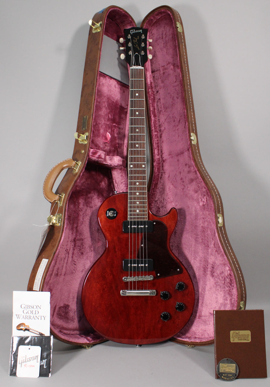 Full view of a dark cherry Les Paul Special with two black P90 pickups, black pickguard, and black control knobs, resting in its open brown case with purple plush lining.